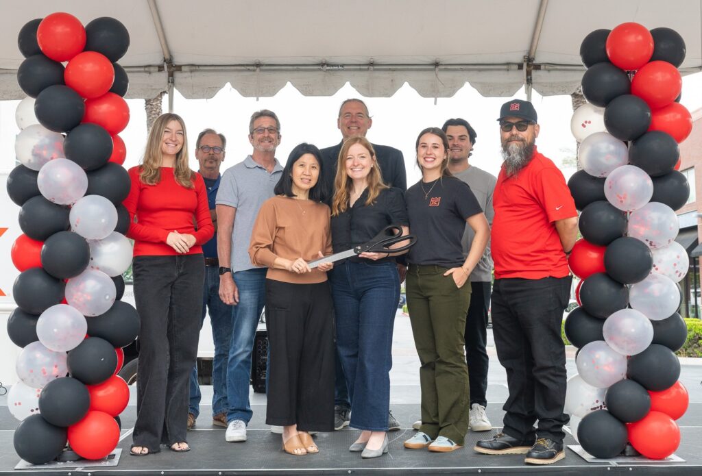 Group of nine adults on a stage during a ribbon-cutting ceremony, two women at the front holding large scissors, framed by red, black, and white balloon arches.